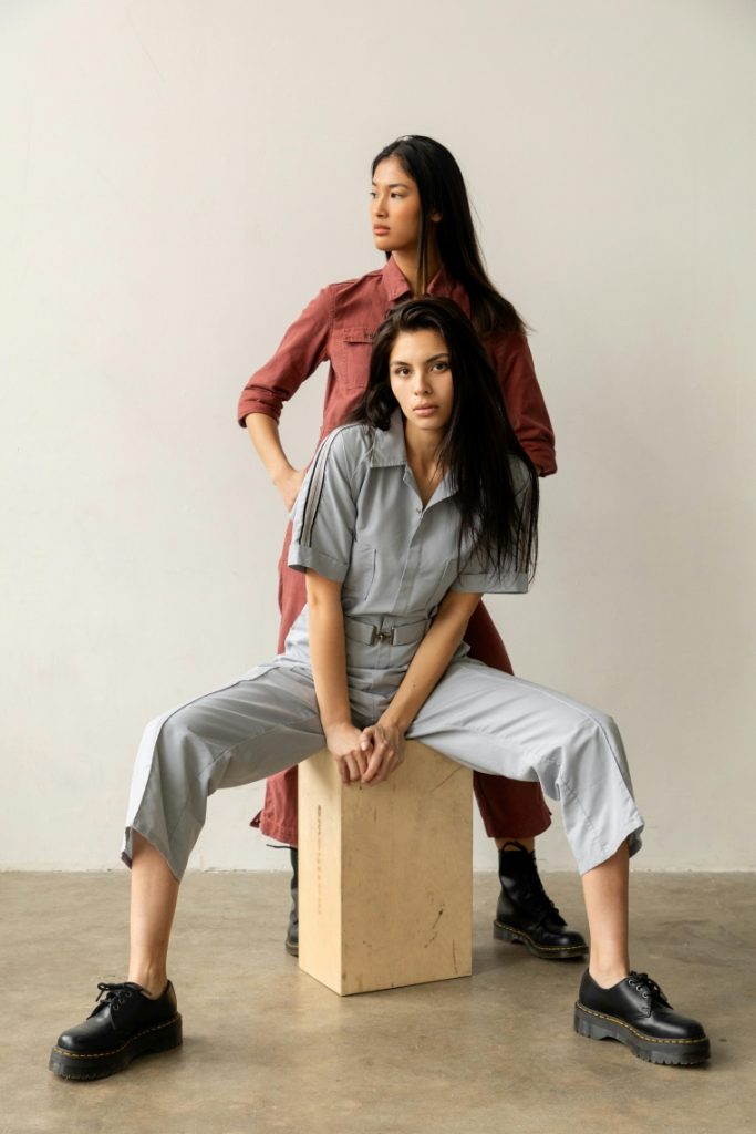 one woman sitting on a box wearing gray jumpsuit for ladies, and another woman standing and wearing red jumpsuit 