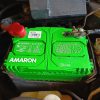 Amaron car battery installed in a vehicle engine compartment, highlighting the brand logo and safety warnings.